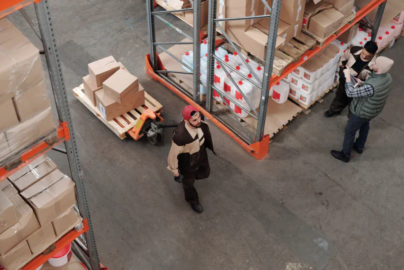 Overhead view of a warehouse worker moving cardboard boxes on a pallet jack between storage racks