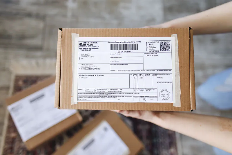 Hands holding a cardboard parcel with a customs declaration form attached to the top