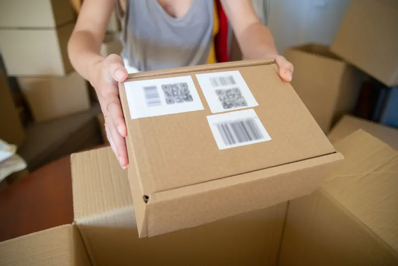 Hands presenting a cardboard box marked with barcode and QR code shipping labels
