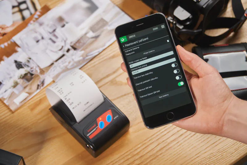 A thermal receipt printer printing a paper slip beside a phone showing printer settings