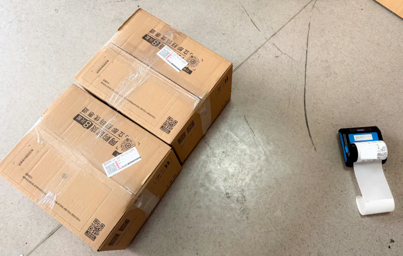 A compact mobile thermal label printer printing a shipping label next to two packed parcels