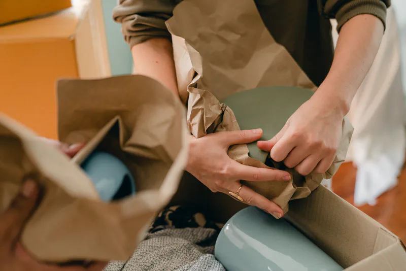 Hands wrapping a green ceramic vase in brown kraft paper before boxing it