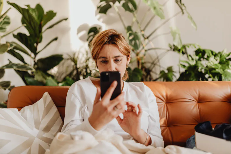 A person relaxing on a couch at home, checking a smartphone with a delivered box nearby