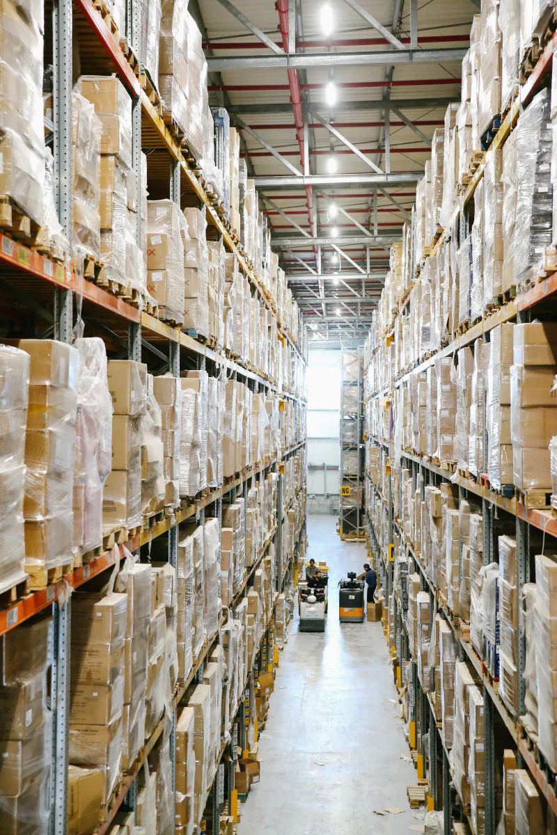 A tall warehouse aisle lined with pallet racks of boxes with workers and a pallet jack in the distance