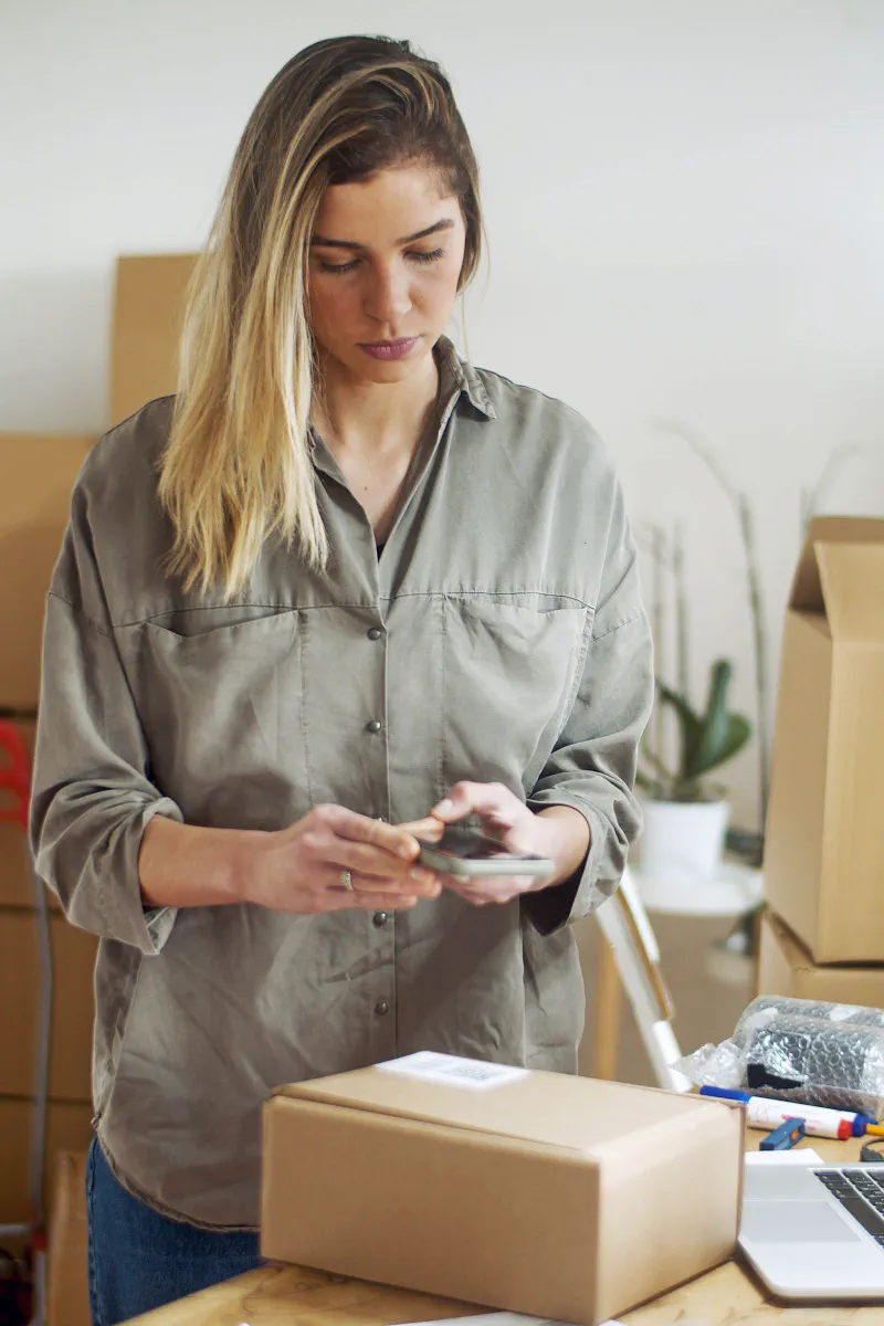 A seller using a smartphone to photograph a packed cardboard parcel at a home fulfillment desk