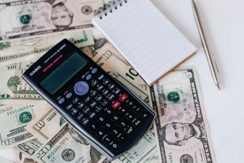 A calculator and a notepad resting on a spread of scattered US dollar bills
