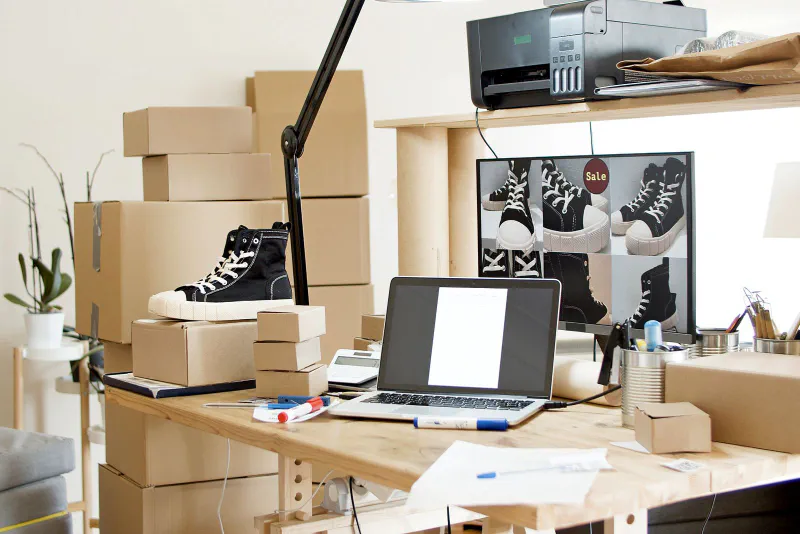 A home shipping workspace with a laptop, a calculator, and stacked cardboard mailing boxes on a wooden desk