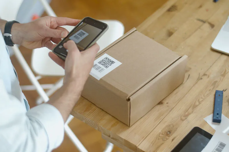 Hands using a smartphone camera to scan the barcode and QR code on a cardboard parcel