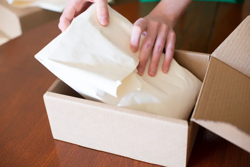 Hands packing an order by placing paper cushioning wrap into a cardboard mailer box on a table